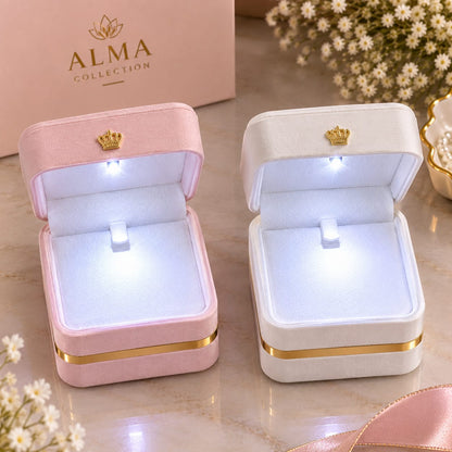 Two velvet ring boxes with built-in LED lights, one pink and one white, both featuring gold trim and a small gold crown emblem on the inside lid. The pink box is closed and shows the ALMA COLLECTION branding on its box.